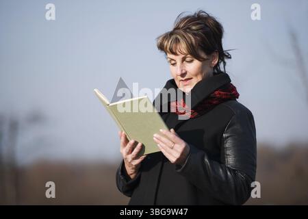 Vista laterale superiore del corpo di una donna di età diversa con una giacca di pelle nera che legge un libro davanti a uno sfondo sfocato Foto Stock
