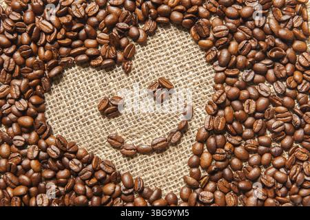 Vista che riempie il formato di una cornice inclinata a forma di cuore composta da chicchi di caffè tostati con un sorriso su un sacco Foto Stock