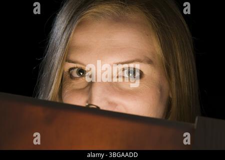 Giovane donna bionda che guarda nella fotocamera da una scatola di legno illuminata internamente che copre il suo volto di fronte a uno sfondo nero Foto Stock