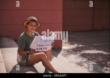 Una donna gode di un momento sereno all'aperto, seduto e leggendo un giornale. Indossa un abbigliamento casual con un cappello, che riflette il relax e il tempo libero nel W Foto Stock