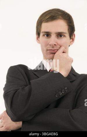 Ripresa della parte superiore del corpo di un giovane uomo d'affari con un abito scuro, braccia piegate e mano sul mento dal basso, guardando seriamente la fotocamera Foto Stock
