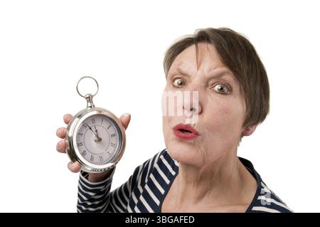 Vista della testa e delle spalle di una donna anziana che tiene un orologio nella mano destra e spaventata con la bocca rotonda guardando la telecamera contro uno sfondo bianco Foto Stock