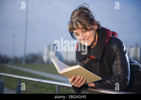 Vista laterale della parte superiore del corpo di una donna matura in una giacca di pelle nera appoggiata su una ringhiera che legge un libro su uno sfondo urbano sfocato Foto Stock