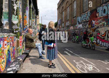 I turisti si fermano a guardare gli artisti di graffiti di Buxton Street, Brick Lane, situato nell'East End di Londra, London Borough of Tower Hamlets, Inghilterra, Regno Unito Foto Stock