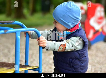 Sorridente bimbo che gioca in un parco giochi, tenendo in mano i bar colorati. Indossato con un berretto e un giubbotto, ama giocare all'aperto con il freddo Foto Stock