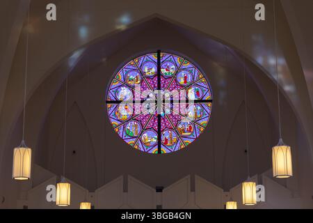 Vista interna di una chiesa caratterizzata da una rosa di vetro colorato circolare illuminata con colori vivaci e lampade sospese. Foto Stock