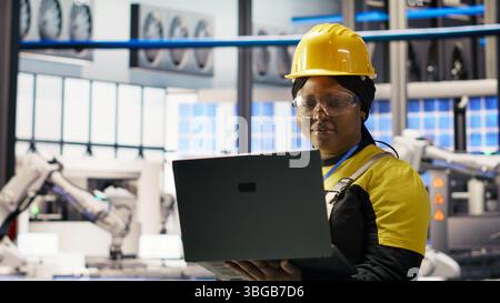 Donna che lavora in un impianto industriale 4,0 e utilizza un notebook, seguendo le procedure di manutenzione. Componente intelligente del personale di fabbrica che analizza i dati sul laptop per prevedere guasti agli ingranaggi, telecamera B. Foto Stock