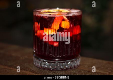 Cocktail rosso con fette di arancia e limone in un bicchiere, servito su un tavolo di legno. Bevanda rinfrescante alla frutta adatta per immagini estive o a tema bar. Foto Stock