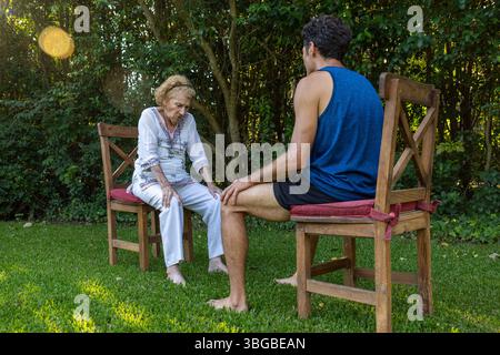 Donna anziana che soffre di dolore al ginocchio che riceve assistenza da un fisioterapista durante una sessione di terapia all'aperto in un tranquillo giardino, prot Foto Stock