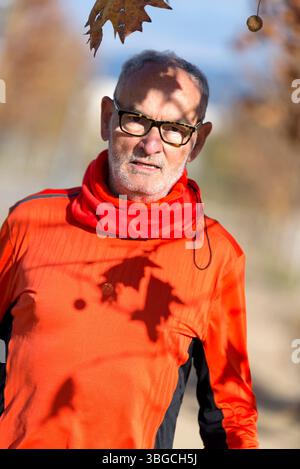 Uomo anziano caucasico con capelli e occhiali grigi, indossa abbigliamento sportivo rosso e sta all'aperto sotto l'ombra degli alberi in una giornata di sole in autunno. Foto Stock