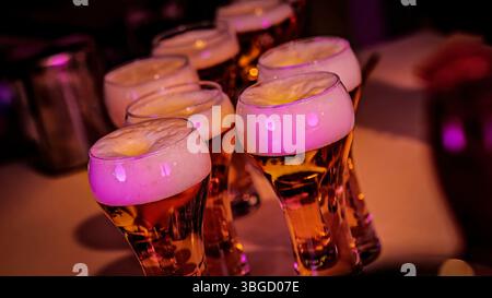 Bicchieri da birra traboccanti con spessa schiuma sul bancone del bar, illuminati da luci rosa, pronti per un brindisi in un pub Foto Stock