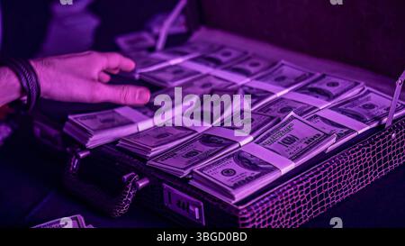 La mano che si avvicinava a pile di banconote da cento dollari erano banchettate e disposte ordinatamente in file all'interno di una valigetta sotto la luce viola Foto Stock