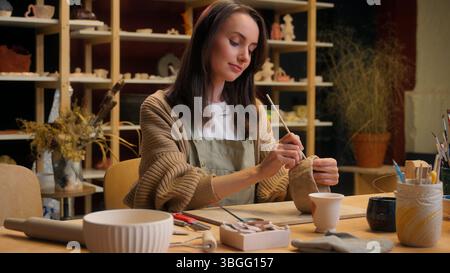 Donna caucasica sorridente decorare stoviglie dipingere argilla in laboratorio di ceramica ragazza artista designer ceramista pittore decoratore arte artigianato hobby Foto Stock