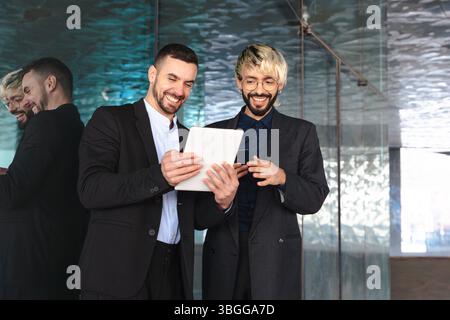 Due uomini d'affari in tuta che usano un tablet insieme e sorridono in un ufficio moderno Foto Stock