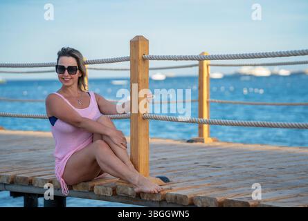Donna seduta su un ponte di legno vicino al mare con un vestito rosa Foto Stock