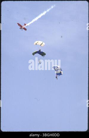 Un biplano arancione si esibisce in uno spettacolo aereo, con tre paracadutisti che scendono dal cielo. I paracadute sono di colore bianco, verde e blu. Foto Stock
