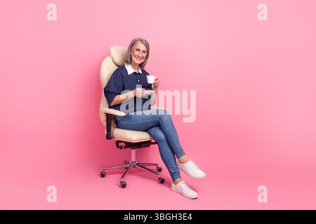 Donna matura in un elegante abbigliamento casual seduta su una sedia con sfondo rosa, godendo del tempo libero e della positività Foto Stock