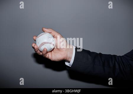 Primo piano su mano di un uomo caucasico di mezza età in tuta che tiene il baseball su sfondo grigio in studio, flash della fotocamera Foto Stock