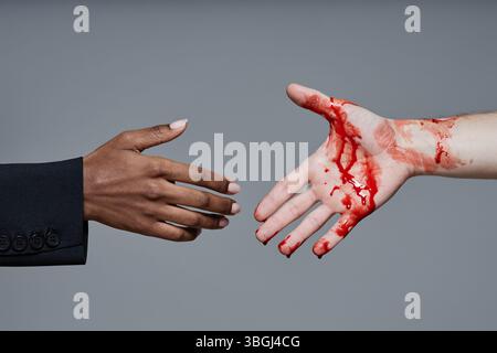 Primo piano sulla mano delle donne nere che si estende verso la mano sanguinosa dell'uomo caucasico che si allunga su sfondo grigio in studio Foto Stock