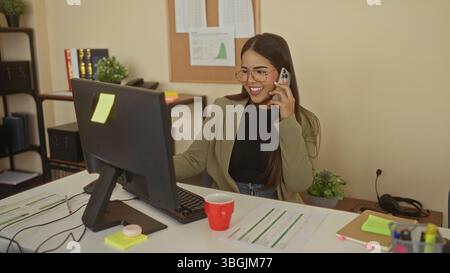 Donna che parla al telefono in ufficio, indossa occhiali e sorride, circondata da monitor e documenti, creando un ambiente di lavoro professionale e coinvolgente Foto Stock