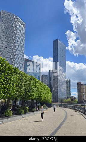 Londra, Regno Unito. 17 aprile 2025, persone che camminano e fanno jogging a Canary Wharf, Londra, con moderni grattacieli e alberi verdi sotto un cielo blu con nuvole Foto Stock