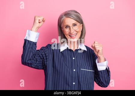 Attraente donna anziana in un abbigliamento elegante che celebra il successo con le mani alzate su un vivace sfondo rosa. Foto Stock