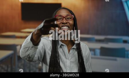 Donna sorridente in classe che indossa occhiali camicia a righe che insegna con gesti espressivi capelli ricci Foto Stock