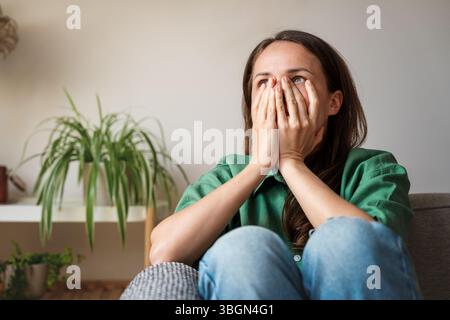 Donna scioccata e stressata con le mani sul viso seduta sul divano di casa Foto Stock