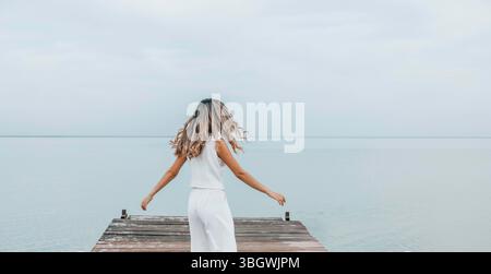 donna irriconoscibile che balla su un molo di legno, capelli lunghi che scorrono liberamente, cielo pallido, circondato da acqua calma, espressione gioiosa in movimento Foto Stock