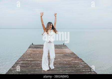 donna che danzava a piedi nudi su un molo di legno, capelli lunghi che scorrevano liberamente sotto un cielo pallido, circondata da acqua calma, espressione gioiosa in movimento Foto Stock
