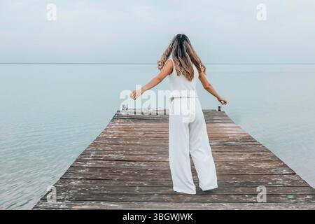 donna irriconoscibile che balla a piedi nudi su un molo di legno, capelli lunghi che scorrono liberamente, cielo pallido, acqua calma, espressione gioiosa in movimento Foto Stock