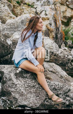 Giovane donna bruna felice con camicia blu, pantaloncini e sandali in denim seduti sulle rocce e guardano con attenzione in lontananza Foto Stock