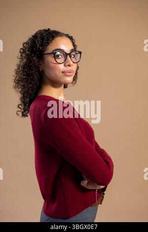 Ripresa in studio laterale di una giovane donna adulta in piedi con le braccia piegate, sorridendo alla telecamera. Indossa occhiali e abbigliamento casual. Foto Stock