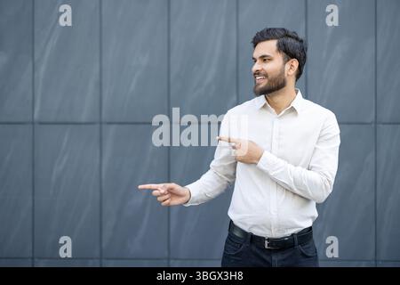 Un uomo sorridente con una camicia bianca punta lateralmente contro lo sfondo di una parete piastrellata grigia. Foto Stock