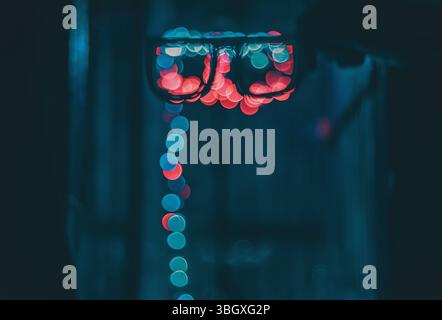 Uno scatto ravvicinato di occhiali che riflettono luci rosse e blu sfocate, creando un effetto bokeh. Le luci sfocate scendono verso il basso. Foto Stock