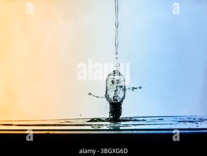 Una fotografia ad alta velocità cattura il momento in cui una goccia d'acqua colpisce una lampadina di vetro, creando un incredibile effetto di spruzzo e ondulazione contro un retrogro gradiente Foto Stock