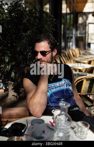 Un uomo elegante con occhiali da sole siede pensiamente ad un tavolo da caffe' all'aperto, godendosi la calda luce del sole durante un momento tranquillo di relax. Foto Stock