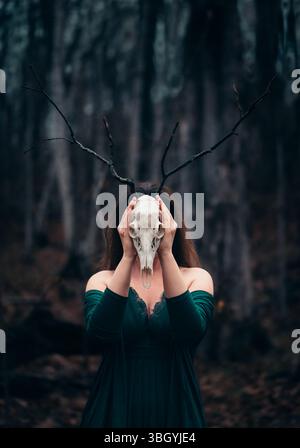 Donna che posa con il teschio di cervo davanti al viso in un ambiente di foresta buia. Foto Stock