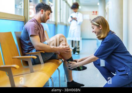 Il giovane seduto in una sala d'attesa dell'ospedale riceve l'assistenza di un infermiere che gli sta mettendo un calzino di compressione sulla gamba Foto Stock