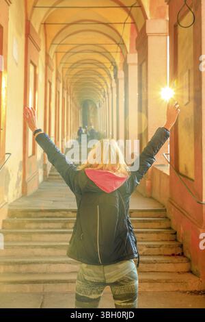 Donna felice con le braccia alzate sotto l'arco di San Luca: L'arco più lungo del mondo che conduce al Santuario di San Luca a Bologna, Italia. Scatto verticale Foto Stock