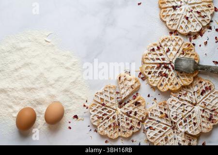 Vista dall'alto di una lastra di marmo bianco con ingredienti per waffle fatti in casa. Accanto alla farina e alle uova potrai vedere waffle pronti. I dolci lo sono Foto Stock