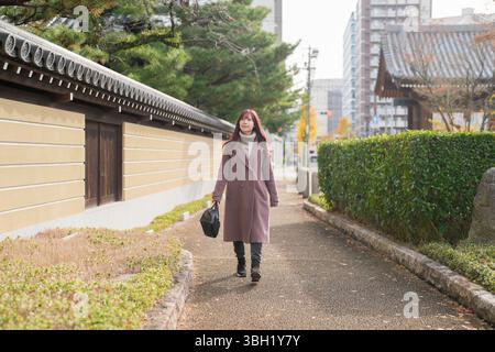 In una fredda mattina di dicembre, una donna giapponese sui vent'anni con lunghi capelli rosa e un trench rosa cammina per una strada tranquilla costeggiata da legno Foto Stock