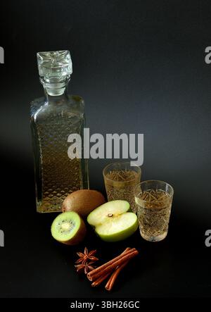 Kiwi e liquore di mele verdi con spezie in vetro cristallo su sfondo nero, accanto a pezzi di frutta matura, cannella e anice. Primo piano. Foto Stock