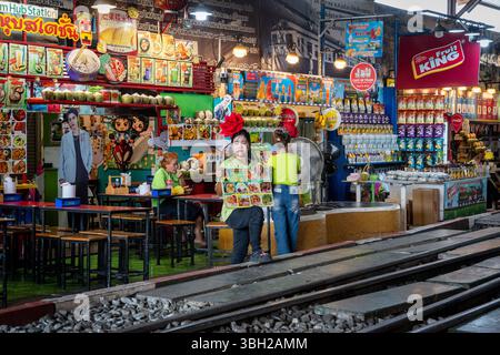 Una vista di una cameriera che cerca di attirare i turisti per mangiare e bere davanti al suo negozio, di fronte all'ufficio della stazione ferroviaria principale presso il mercato ferroviario di Maeklong a Samut Songkhram, Thailandia. Il mercato ferroviario di Maeklong, comunemente chiamato mercato di Siang Tai (rischio di vita), è un mercato locale affollato e popolare luogo turistico nella provincia di Samut Songkhram, in Thailandia. I treni passano più volte al giorno e i venditori ritirano rapidamente i loro ombrelli e le loro esposizioni poco prima dell'arrivo del treno, quindi restituiscono tutto dopo. Il mercato vende prodotti freschi, pesce, spuntini, abbigliamento e souvenir come un Foto Stock