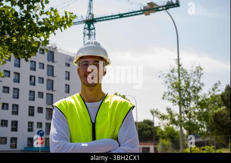 Lavoratore edile professionista con casco di sicurezza e giubbotto riflettente sorridente in un cantiere con i progressi di costruzione in Foto Stock