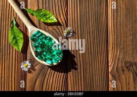 Cucchiaio di legno con capsule verdi e foglie fresche su una superficie di legno. Primo piano di un cucchiaio di legno riempito di capsule verdi, accompagnato da verde Foto Stock