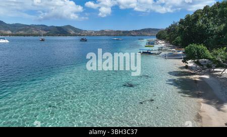 L'acqua limpida e poco profonda rivela il fondale sabbioso lungo una spiaggia tranquilla punteggiata di barche, con montagne panoramiche che si innalzano sullo sfondo sotto il cielo soleggiato Foto Stock