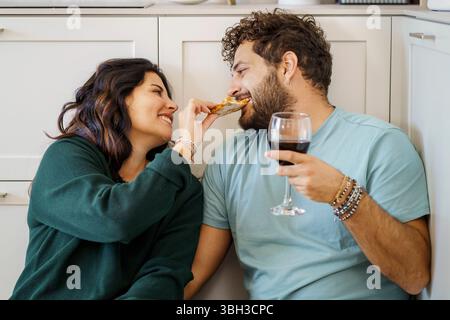 Una giovane coppia affettuosa si gode un pasto informale e romantico a casa, seduti sul pavimento della cucina. La donna nutre giocosamente al suo partner una fetta di pi Foto Stock