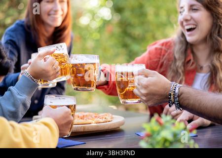 Amici felici che ridono e brindano con tazze da birra su una pizza a un tavolo all'aperto. Una gioiosa scena di festeggiamenti, amicizia, e gustare il cibo e. Foto Stock