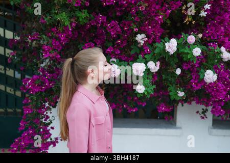 Donna sorridente in giacca rosa che posa accanto alle bouganville magenta in fiore e alle rose bianche. Moda floreale primaverile e bellezza naturale Foto Stock